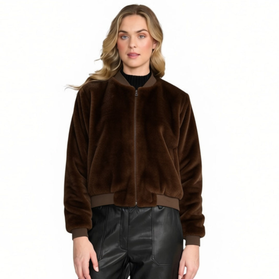 A woman modelling a dark brown faux-fur cropped bomber jacket with a front zipper, ribbed collar, cuffs, and hem, paired with black leather pants against a plain white background.
