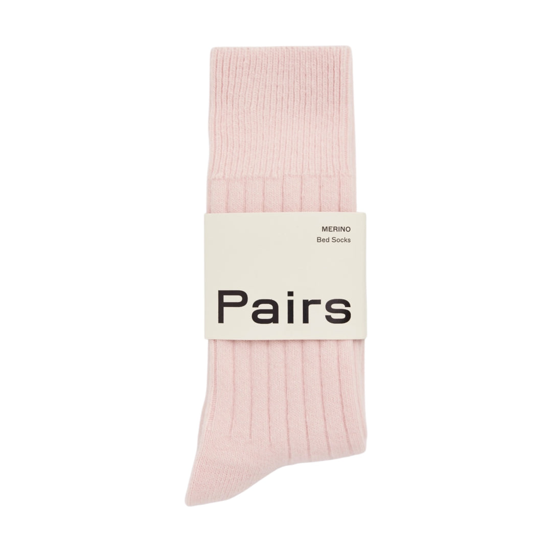 A pair of light pink ribbed bed socks bundled together with a white paper band that reads 'Pairs' and 'MERINO Bed Socks' in black text, set against a plain white background.